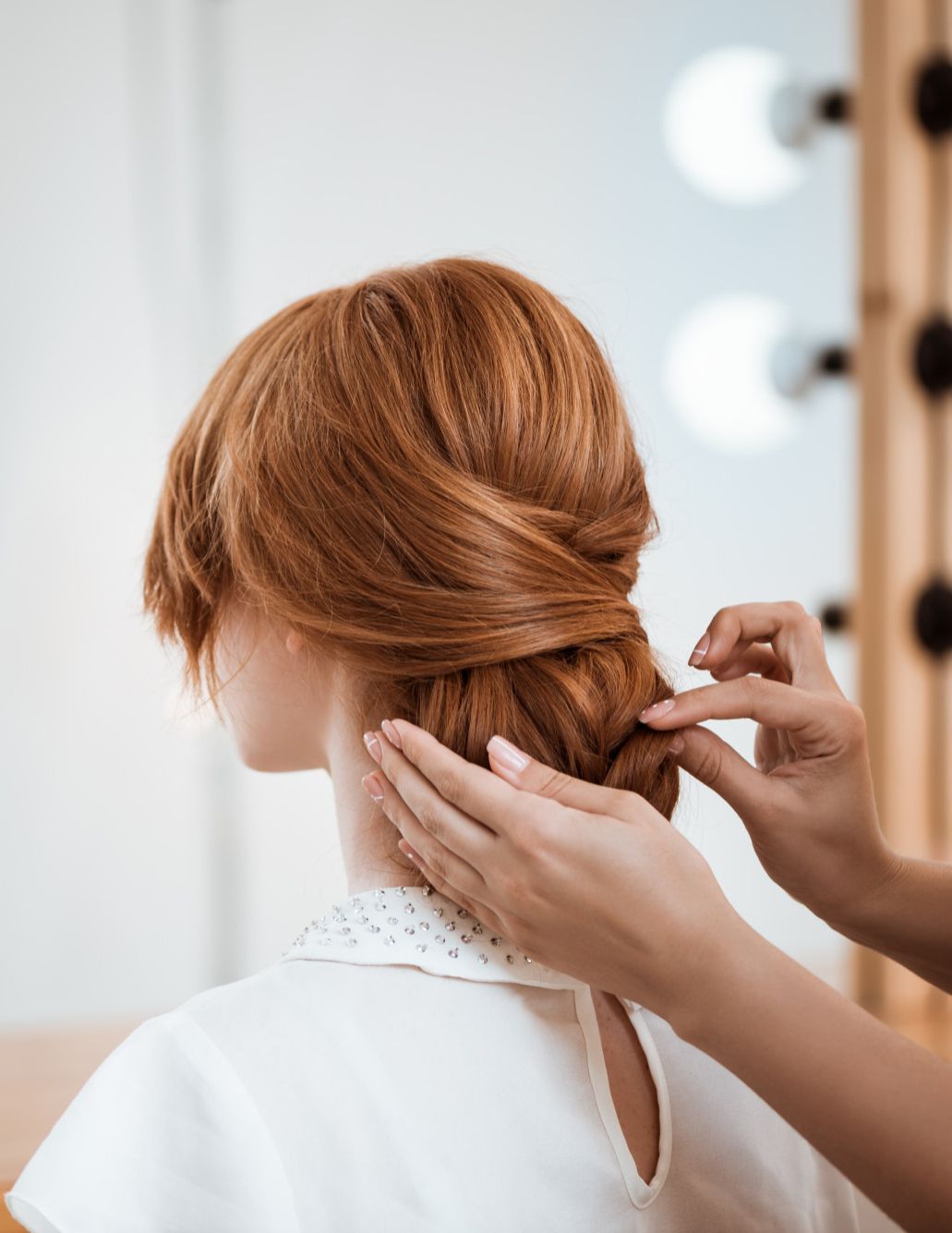 Woman having her red hair styled into a low bun, hands arranging strands, in front of a mirror.