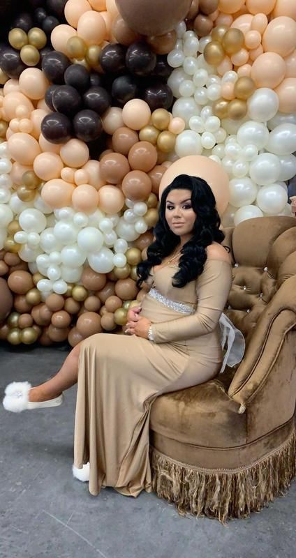 A pregnant woman is sitting on a chair in front of a wall of balloons.
