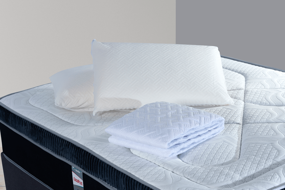 Colchón con almohadas blancas, toalla doblada y cojín decorativo, dentro de una habitación.