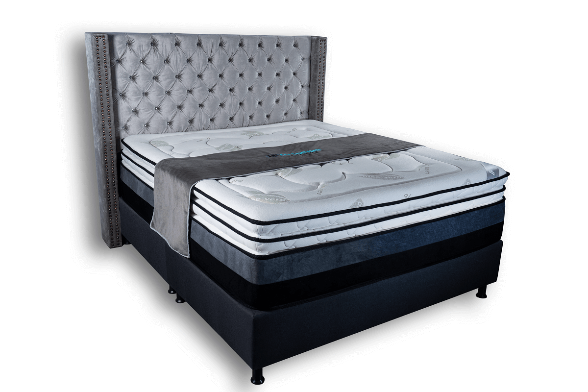 Cama tapizada en gris con cabecera capitoné, colchón de capas y base oscura, sobre fondo blanco.