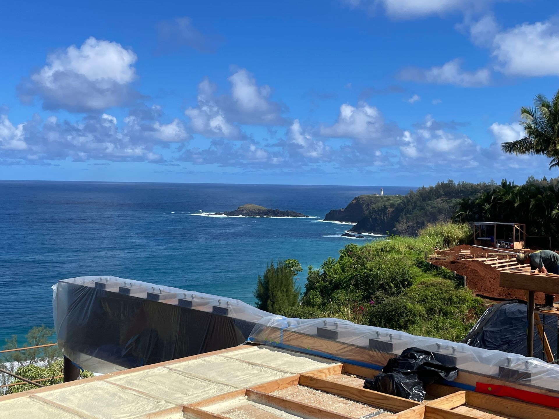 Construction site overlooking ocean; blue sky, white clouds, green trees.