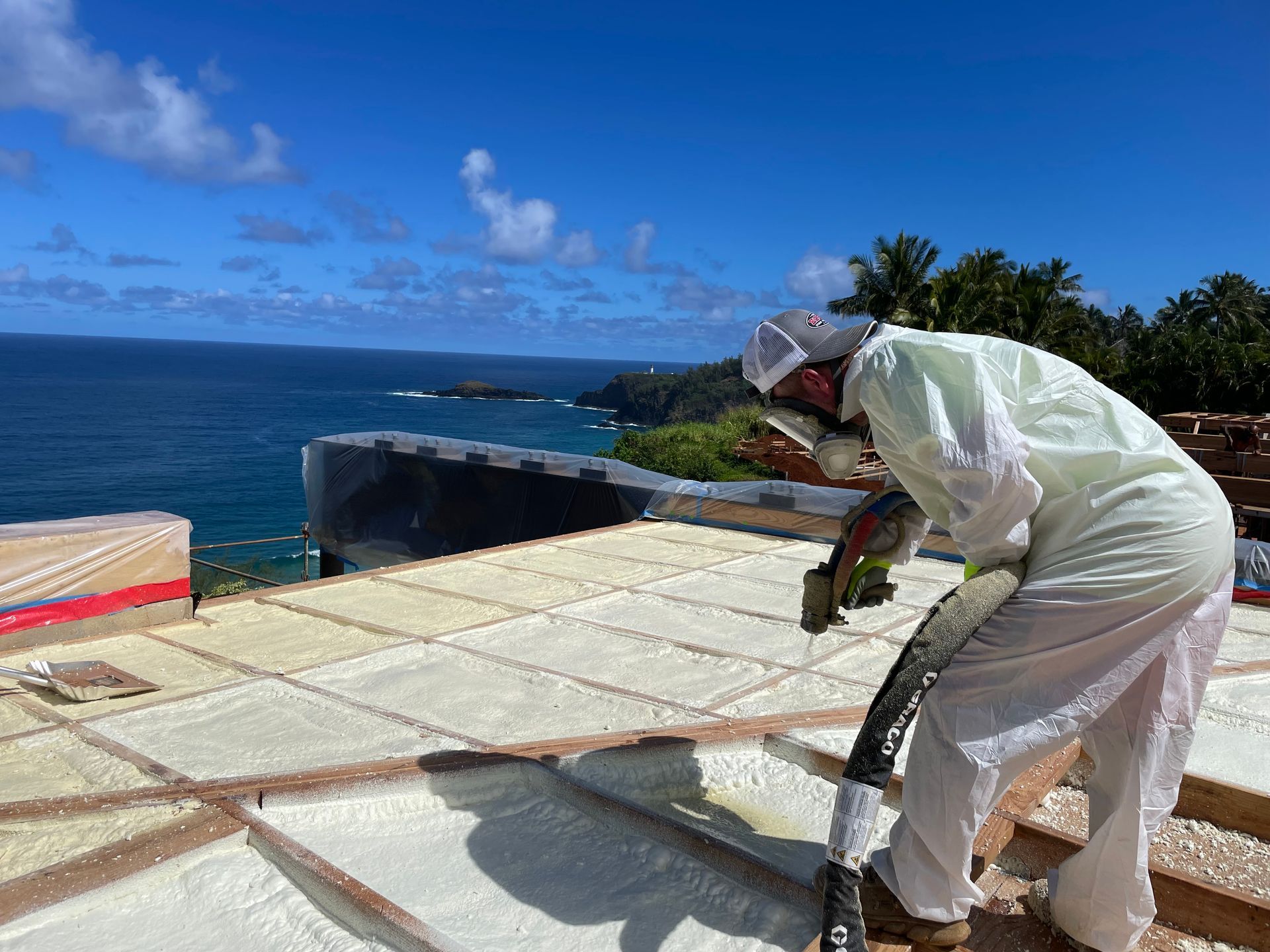 Man in protective suit spraying foam insulation on a roof overlooking the ocean.