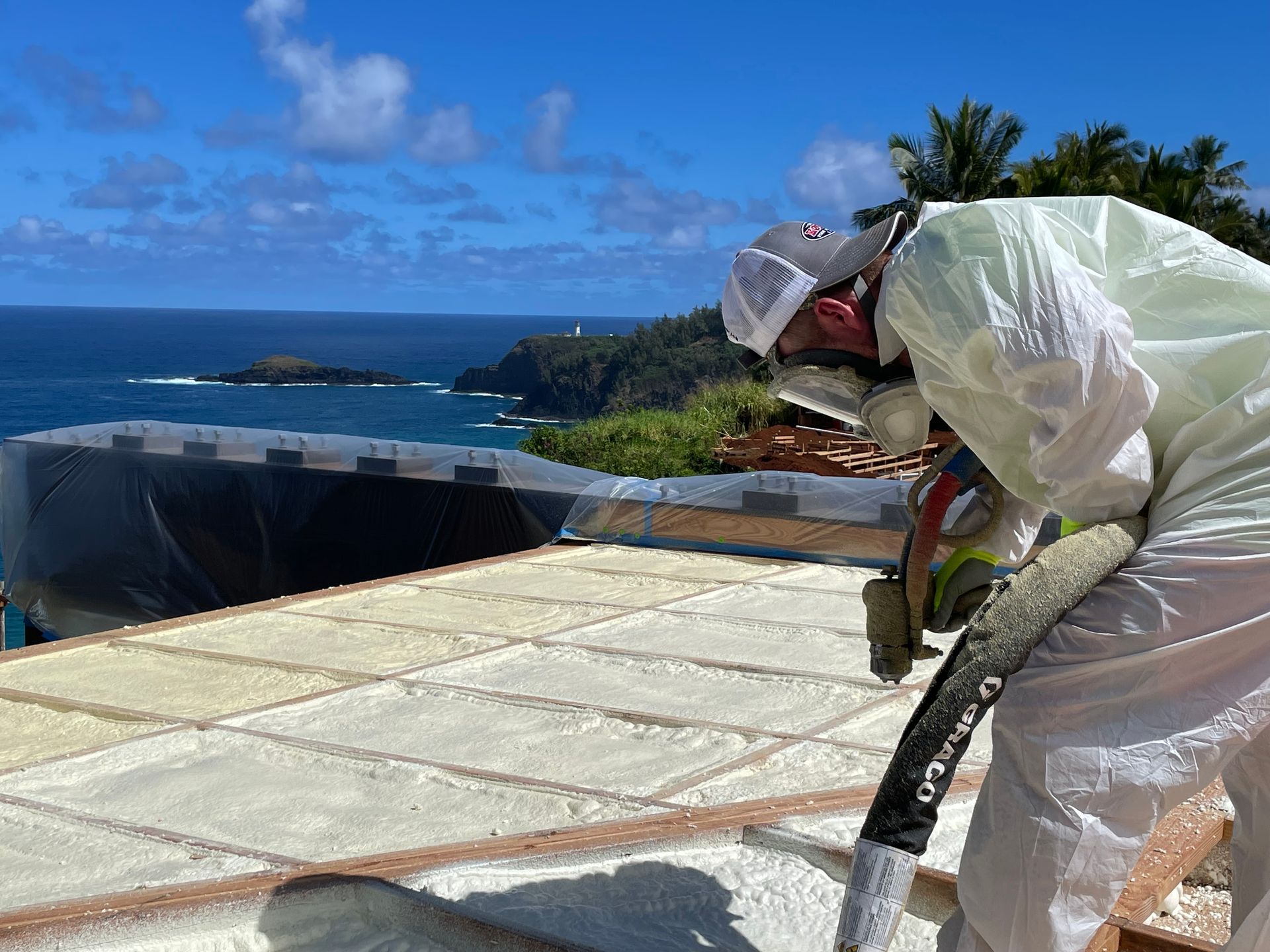 Person in protective suit spraying foam insulation on a roof with ocean view.