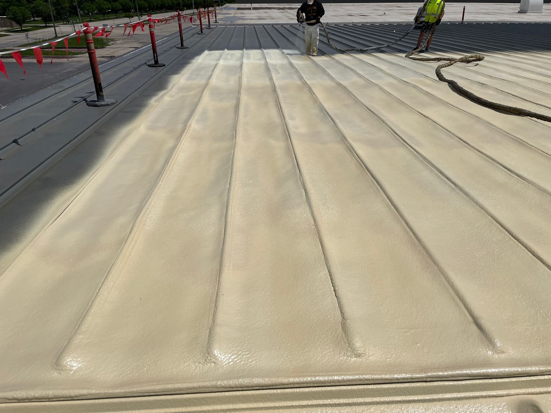 Workers applying tan foam insulation to a corrugated roof, outdoors on a sunny day.