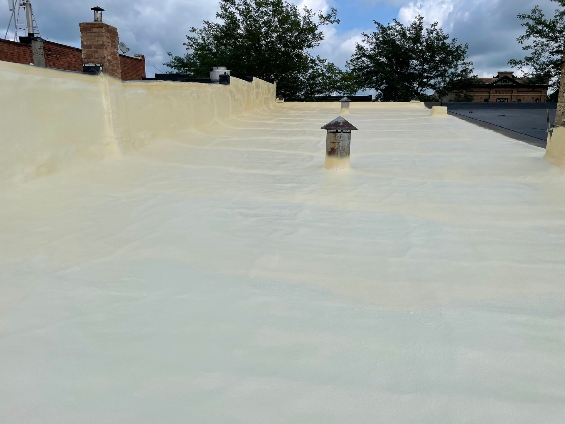 Flat roof covered in light beige spray foam insulation, with chimneys and trees visible.