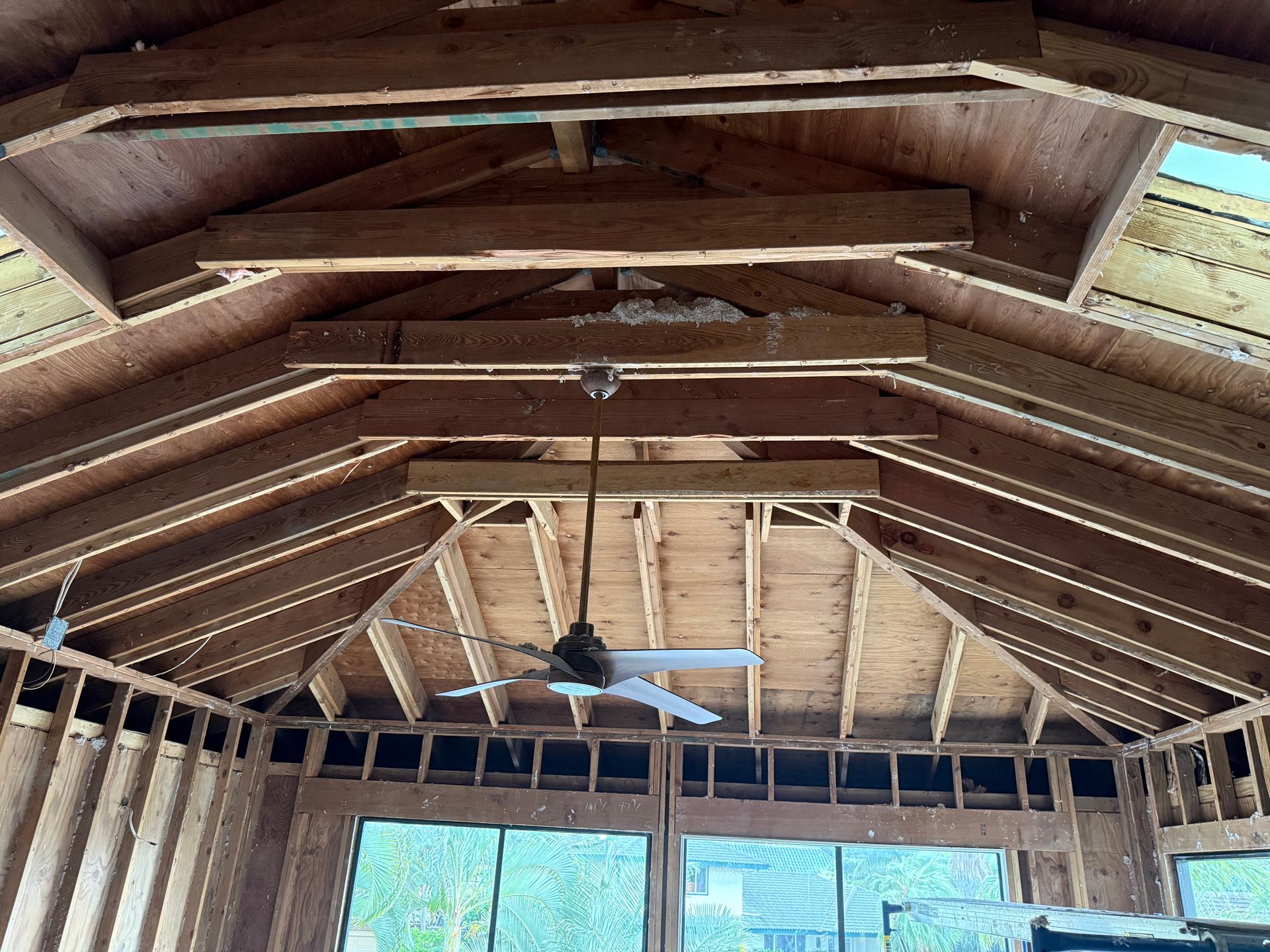 Wooden ceiling and exposed beams inside a structure with a ceiling fan and windows.