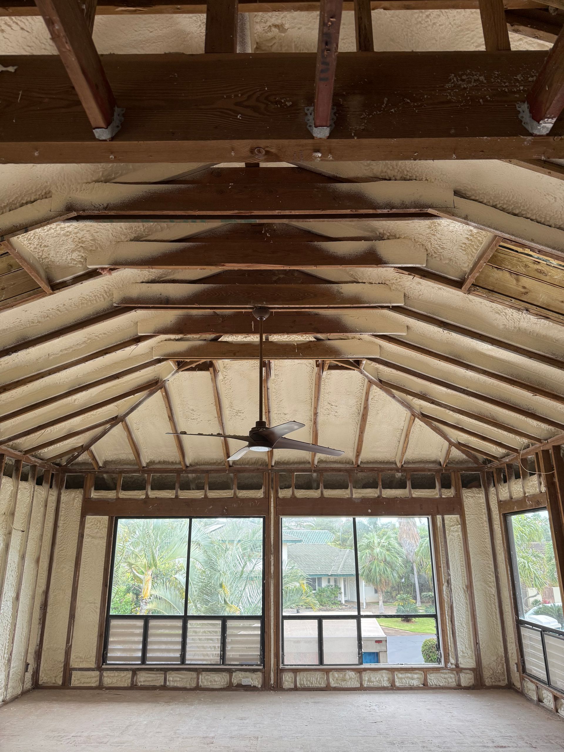 Interior view of a room with spray foam insulation on the walls and ceiling, exposed wooden beams and a ceiling fan.