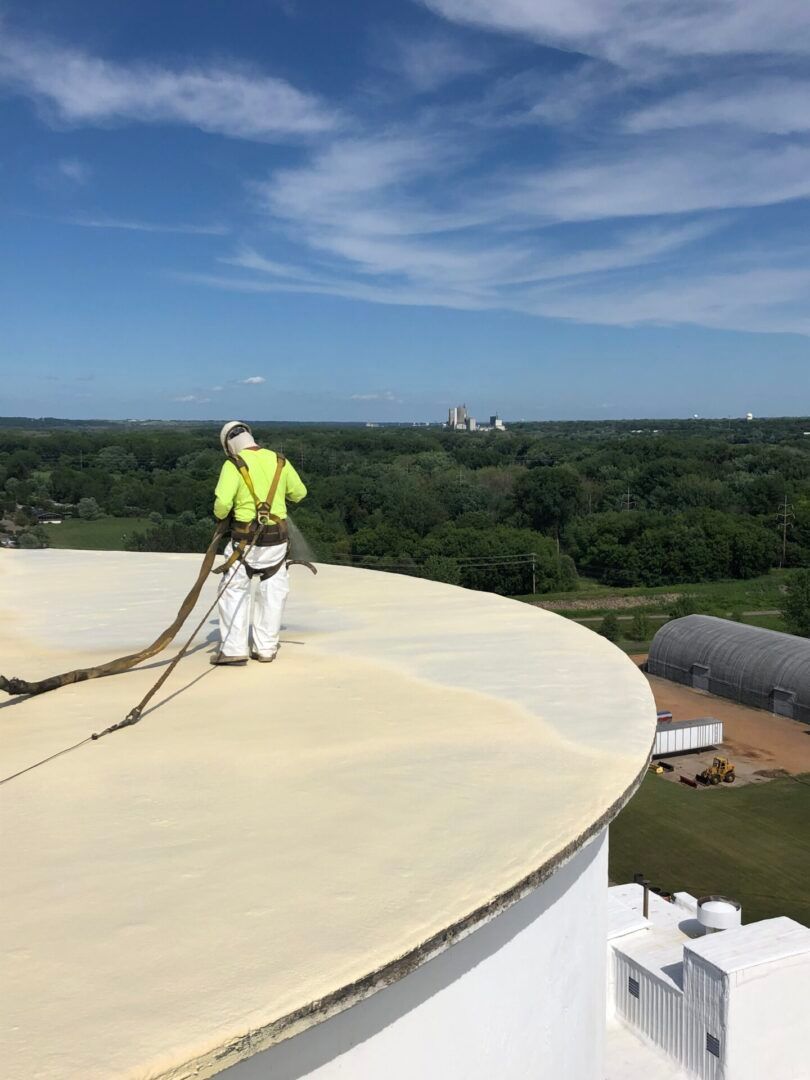 Worker in neon safety gear stands on a large roof under a blue sky, overlooking a wooded landscape.