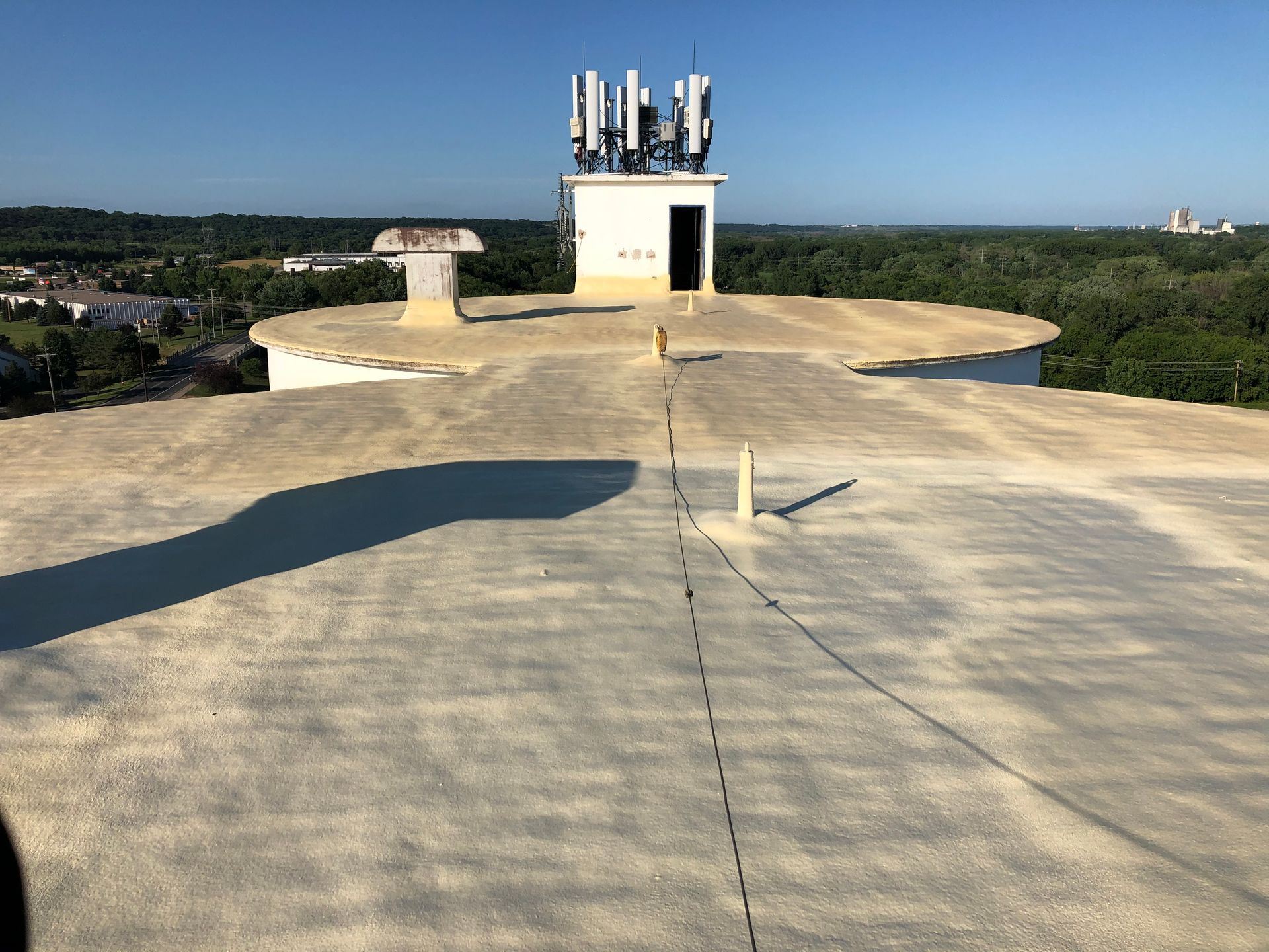 Flat, concrete rooftop with communication equipment on a small building in the center, surrounded by trees and a clear sky.