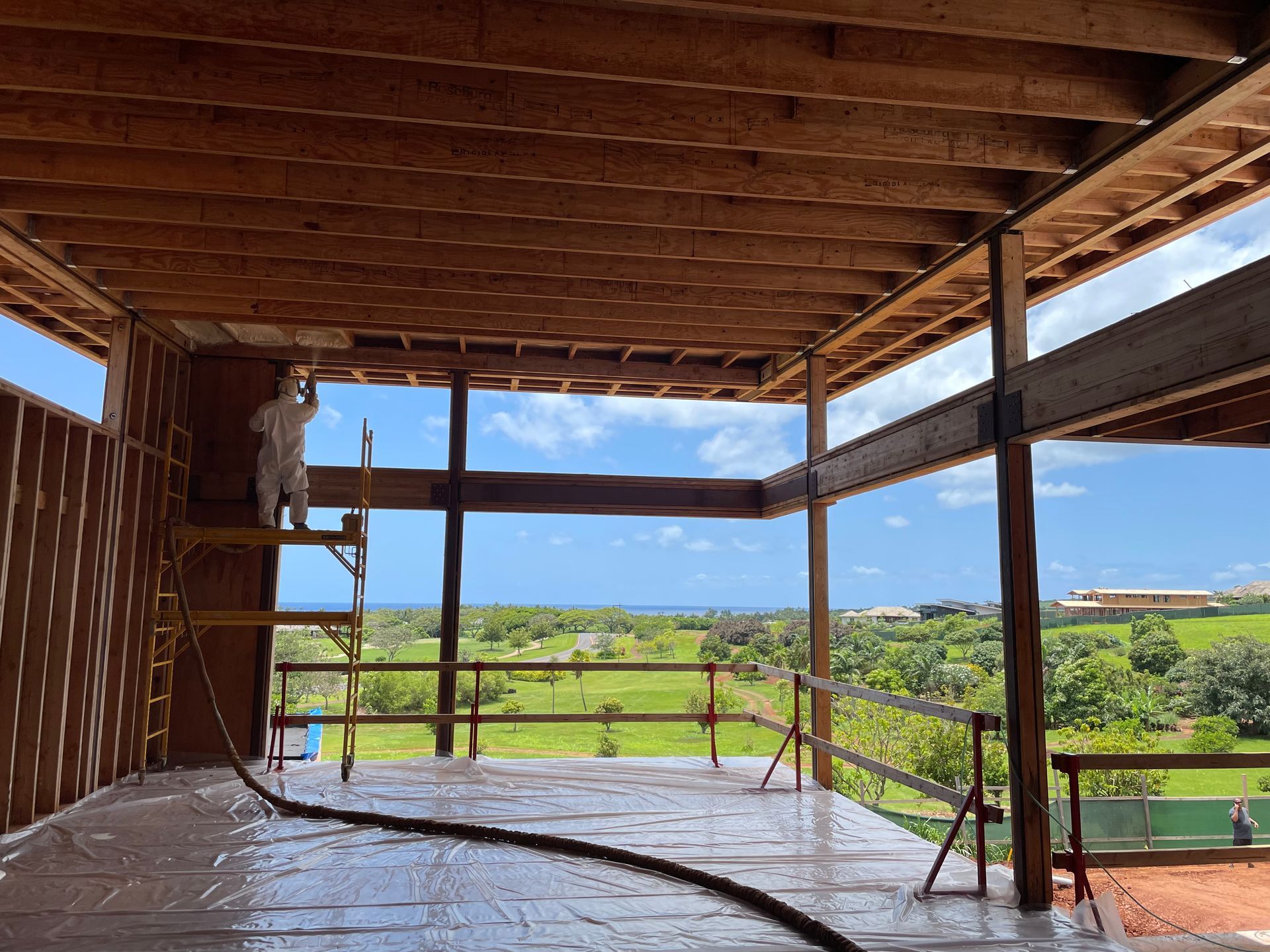 Construction site: person spraying insulation inside a wooden-framed structure, overlooking a green landscape and blue sky.
