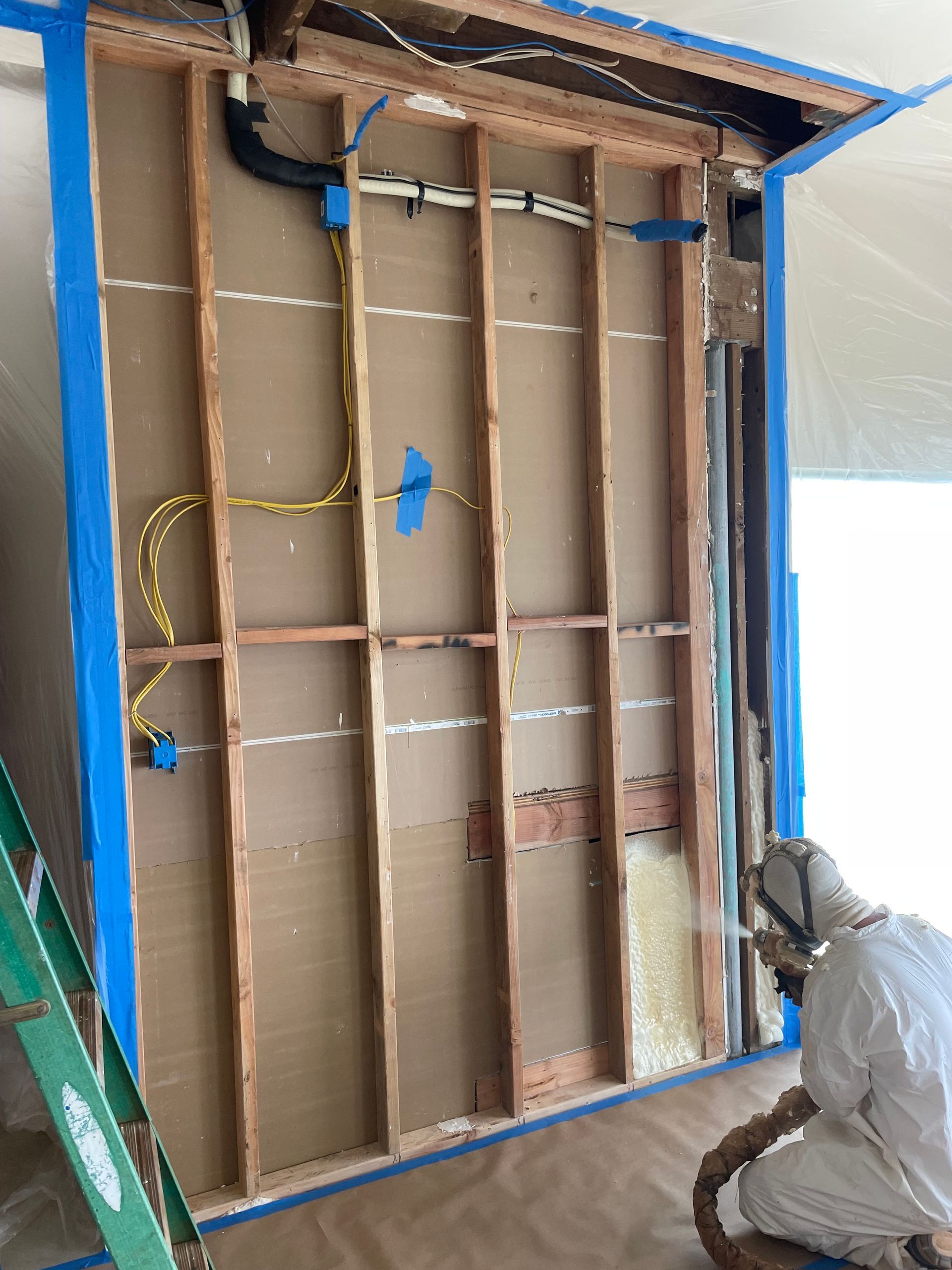 Man in protective suit spraying insulation into exposed wall. Blue tape surrounds the opening.