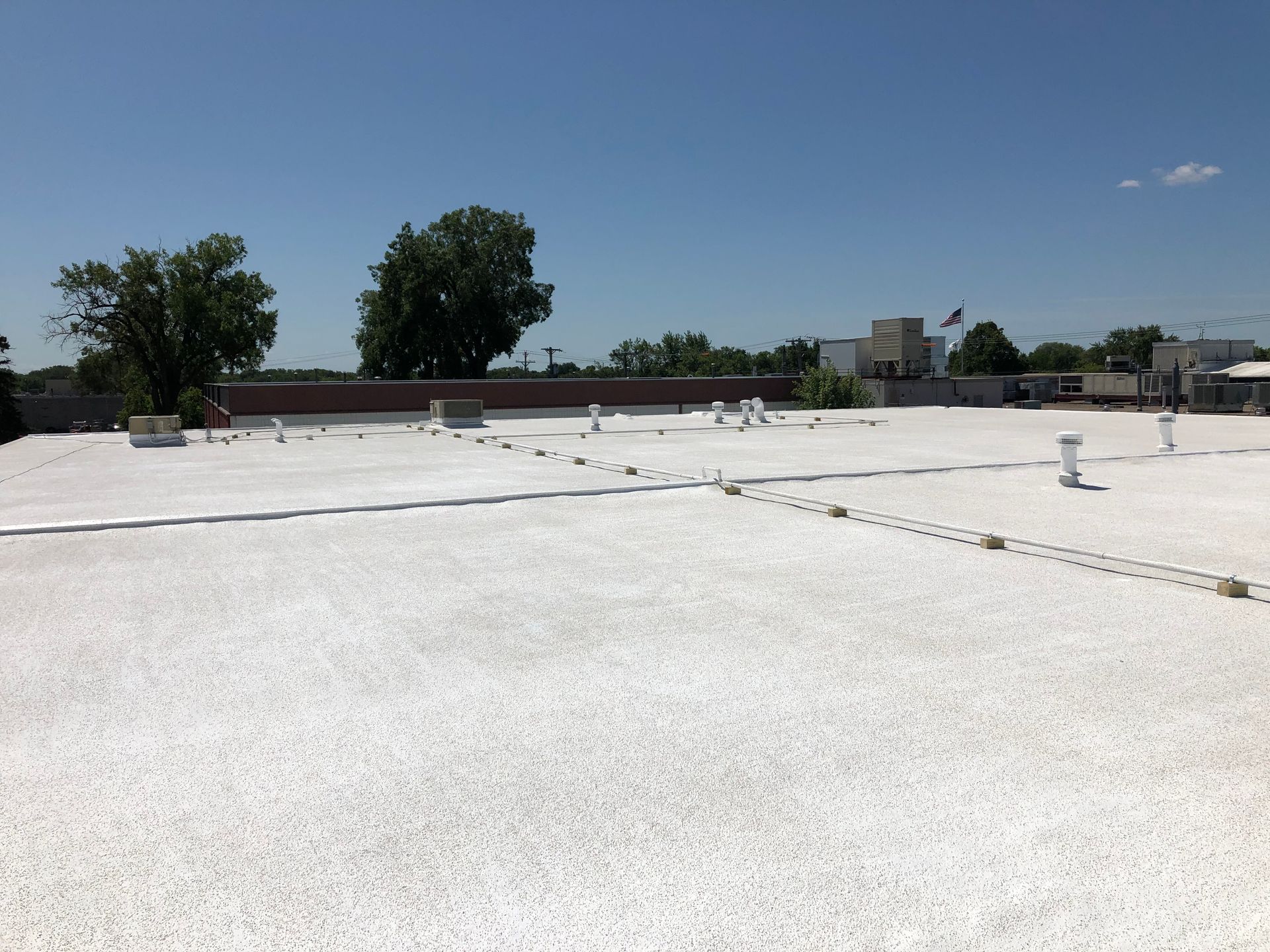 Flat white commercial roof with vents and a few trees against a clear blue sky.