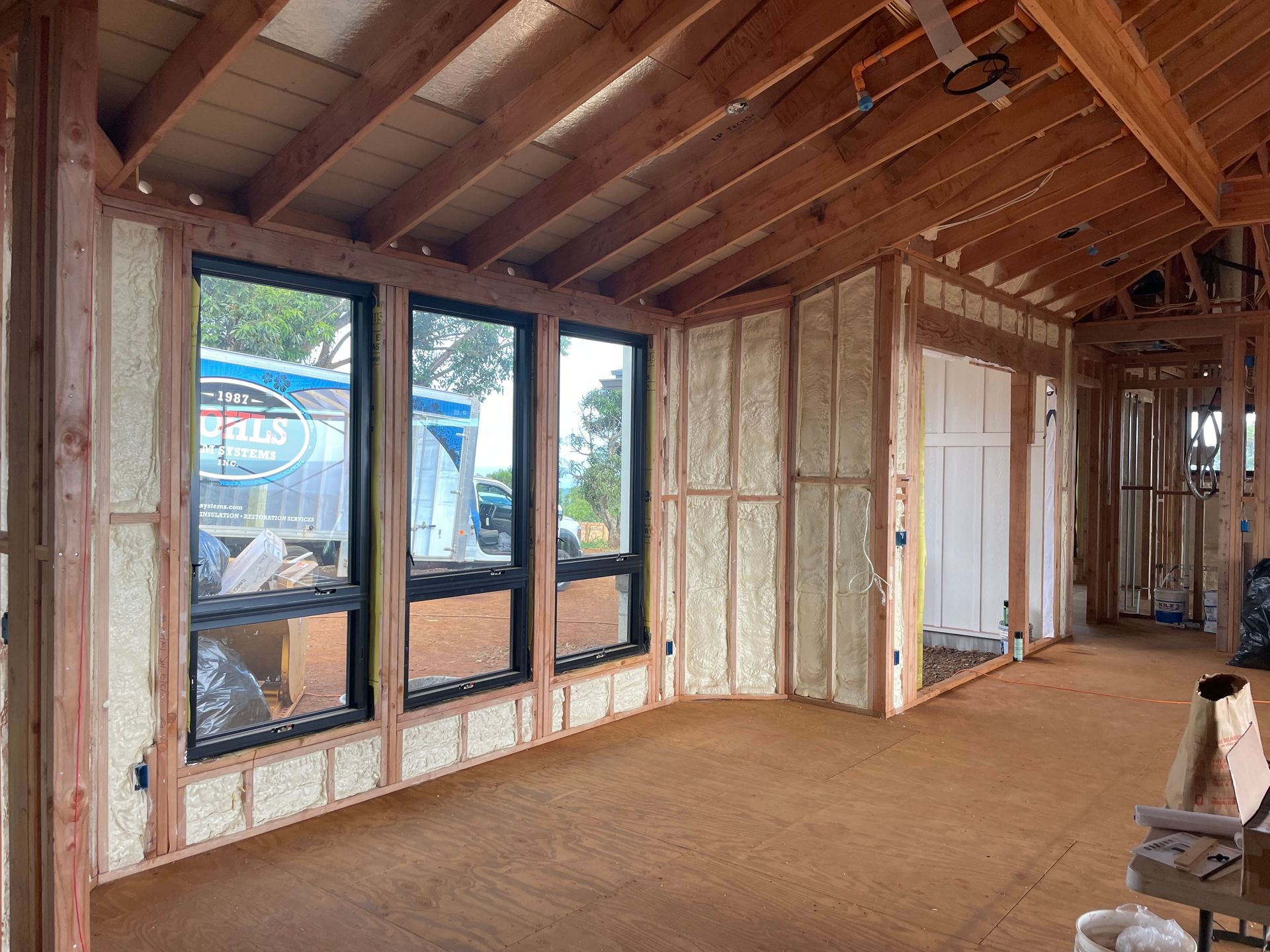 Interior of a room under construction, with spray foam insulation between wooden studs, windows, and exposed ceiling beams.