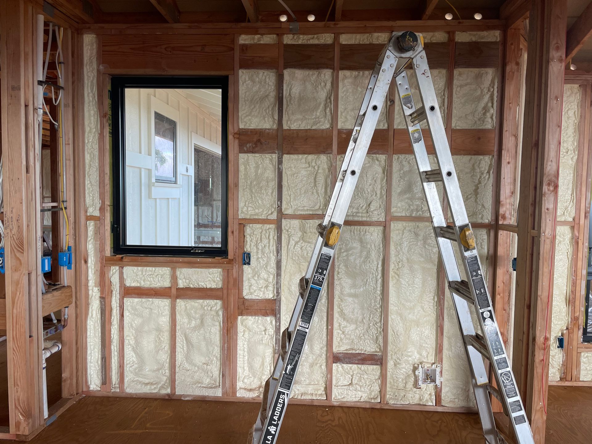 Interior wall under construction, insulation between wooden studs, window with black frame, ladder propped against wall.