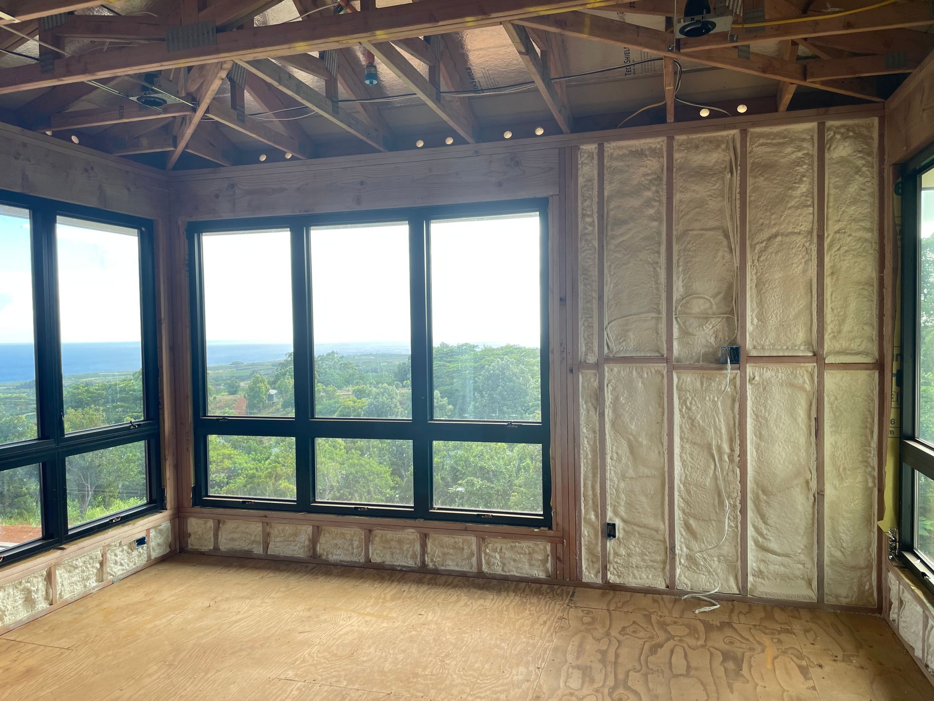Interior of a room under construction, with insulated walls, large windows, and a view of trees.