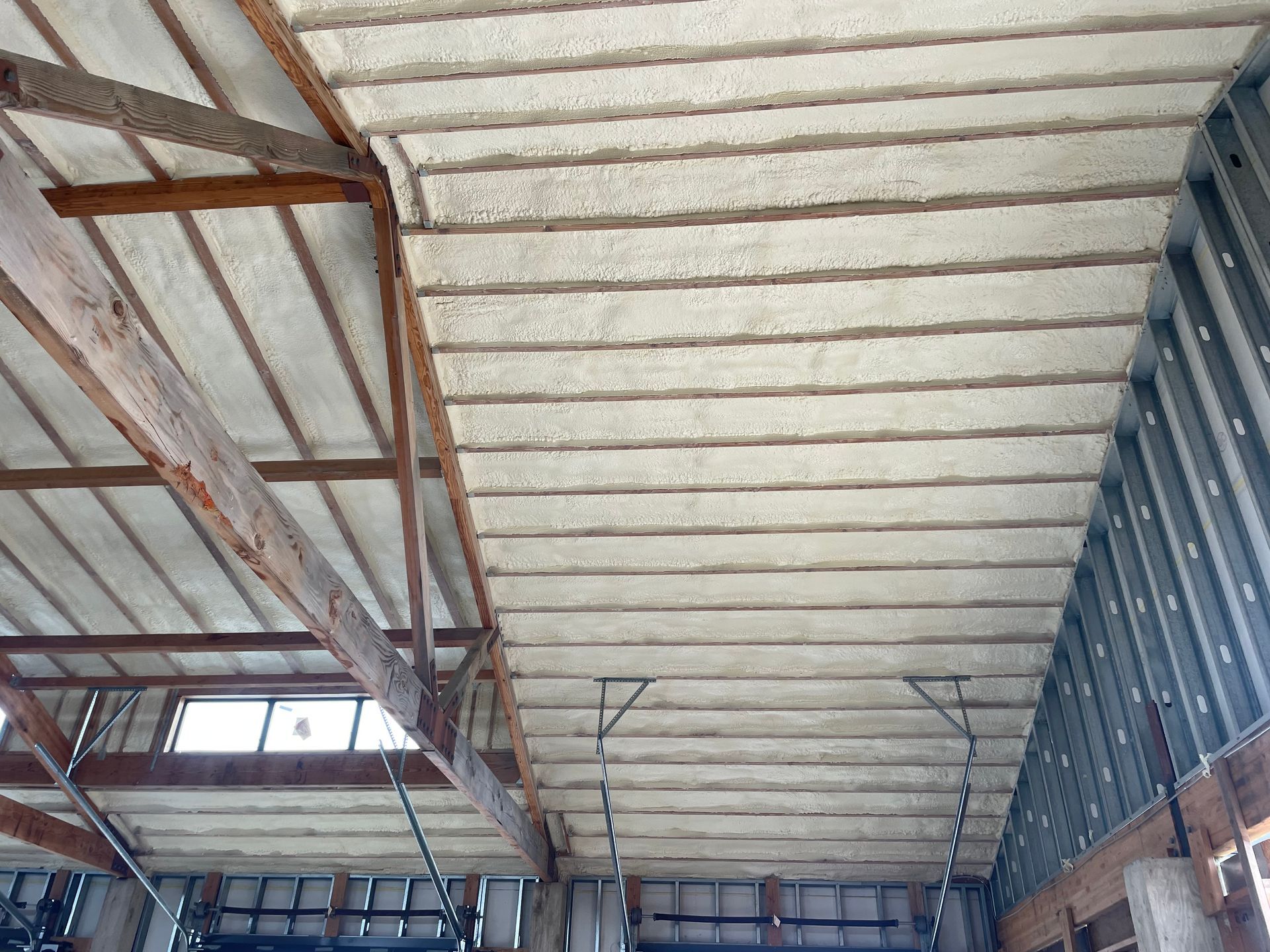 Interior view of a building's ceiling, with wooden beams and white insulation between them, metal framing on the right.