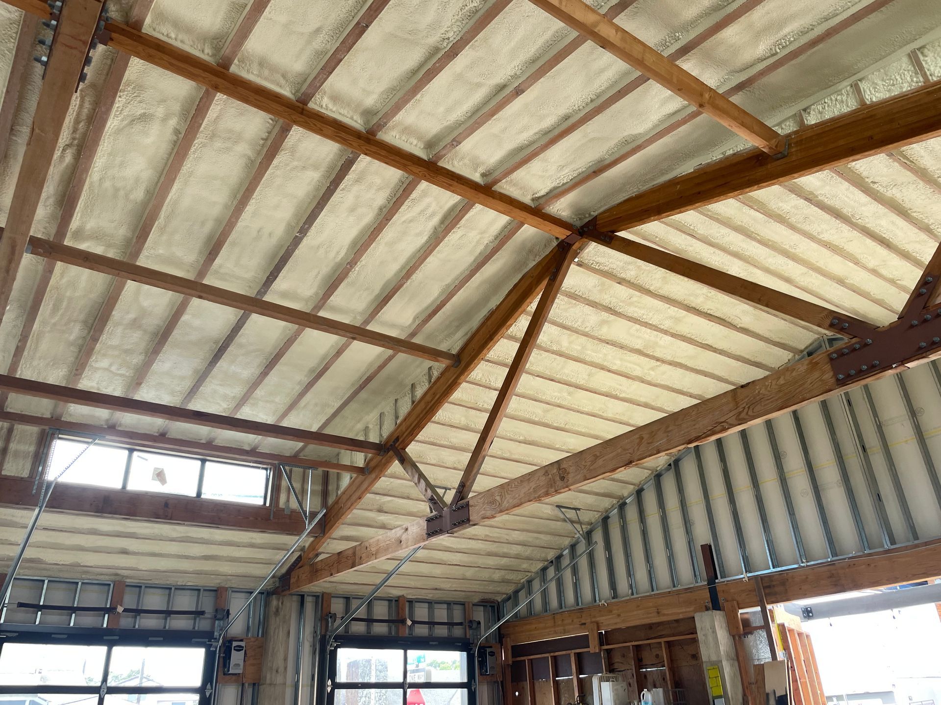 Interior view of a building's roof with spray foam insulation between wooden beams and corrugated metal.