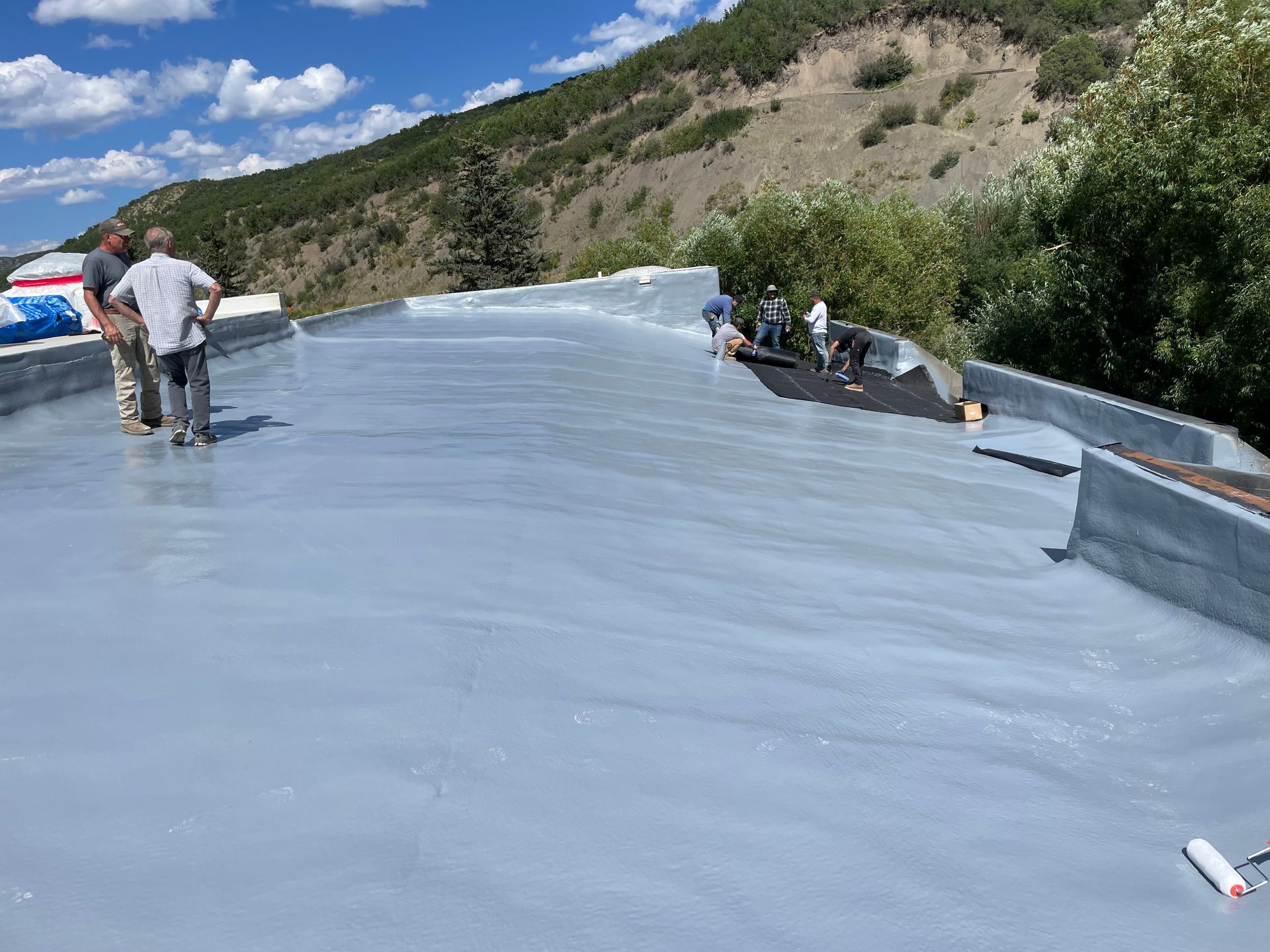 Workers applying gray sealant to a flat roof, with a hillside in the background.