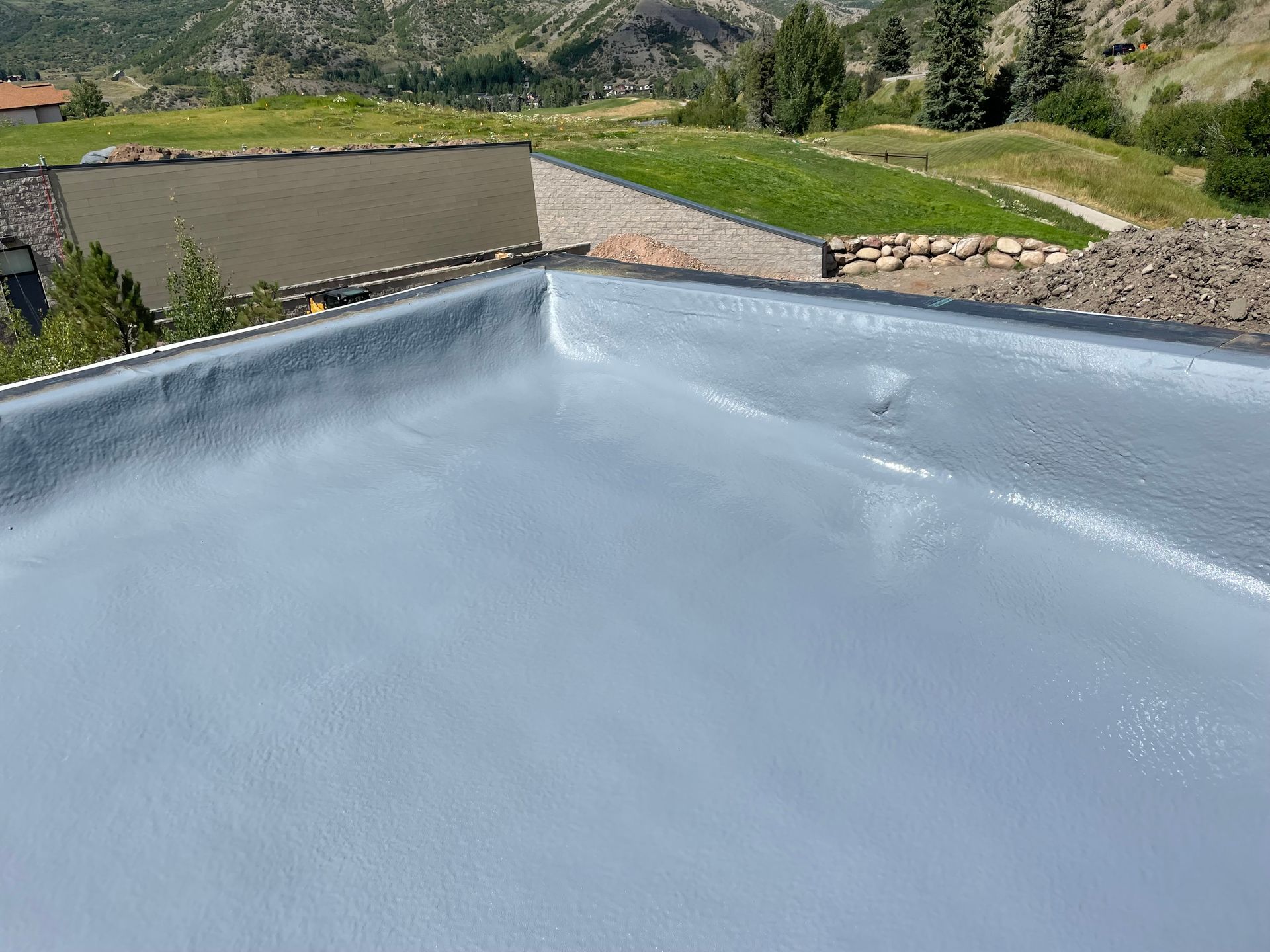 Newly coated flat roof, light gray, with edge detail and background of hillside and building.