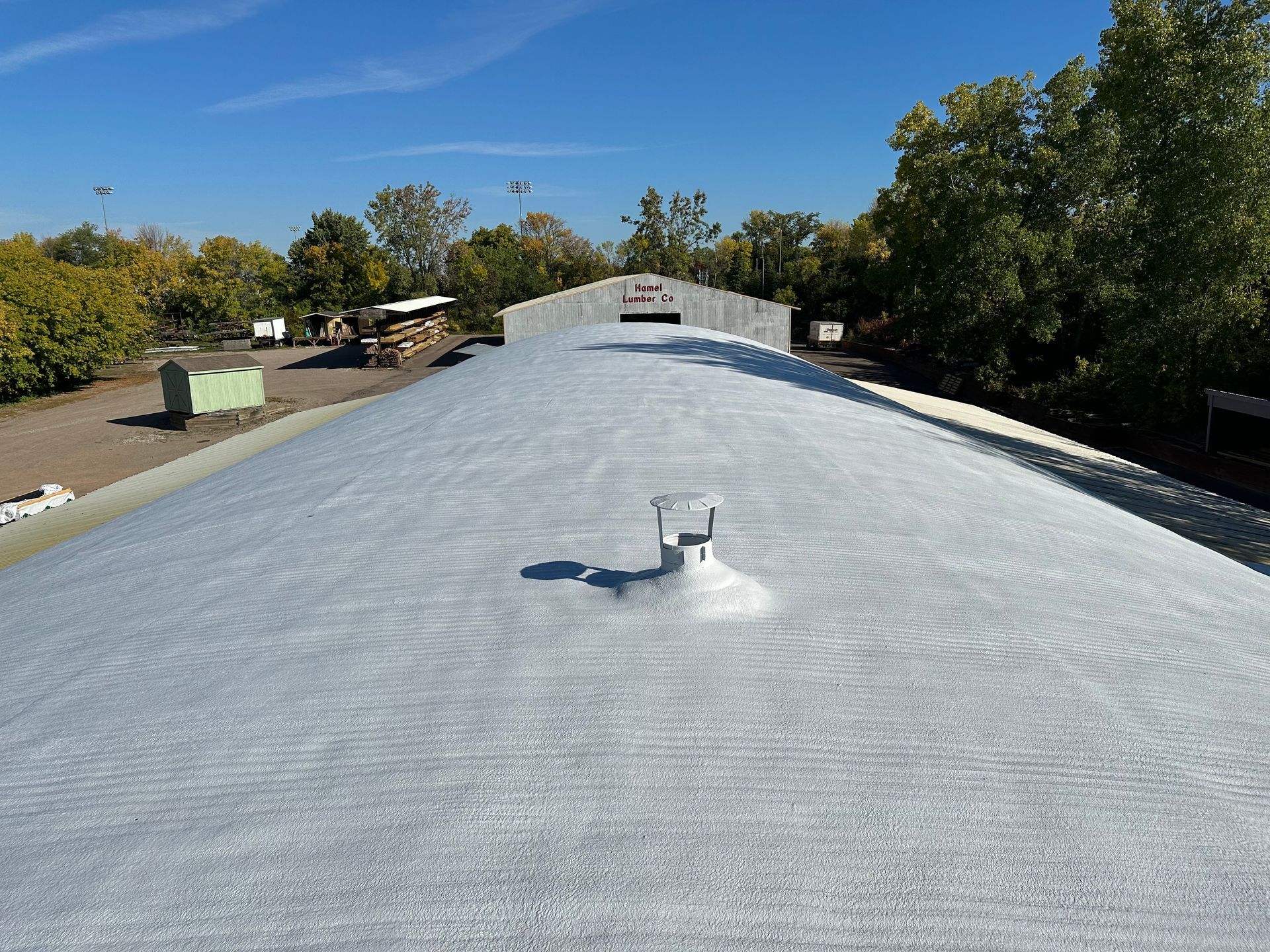 Gray metal roof on a building, a vent pipe, and trees in the background on a sunny day.
