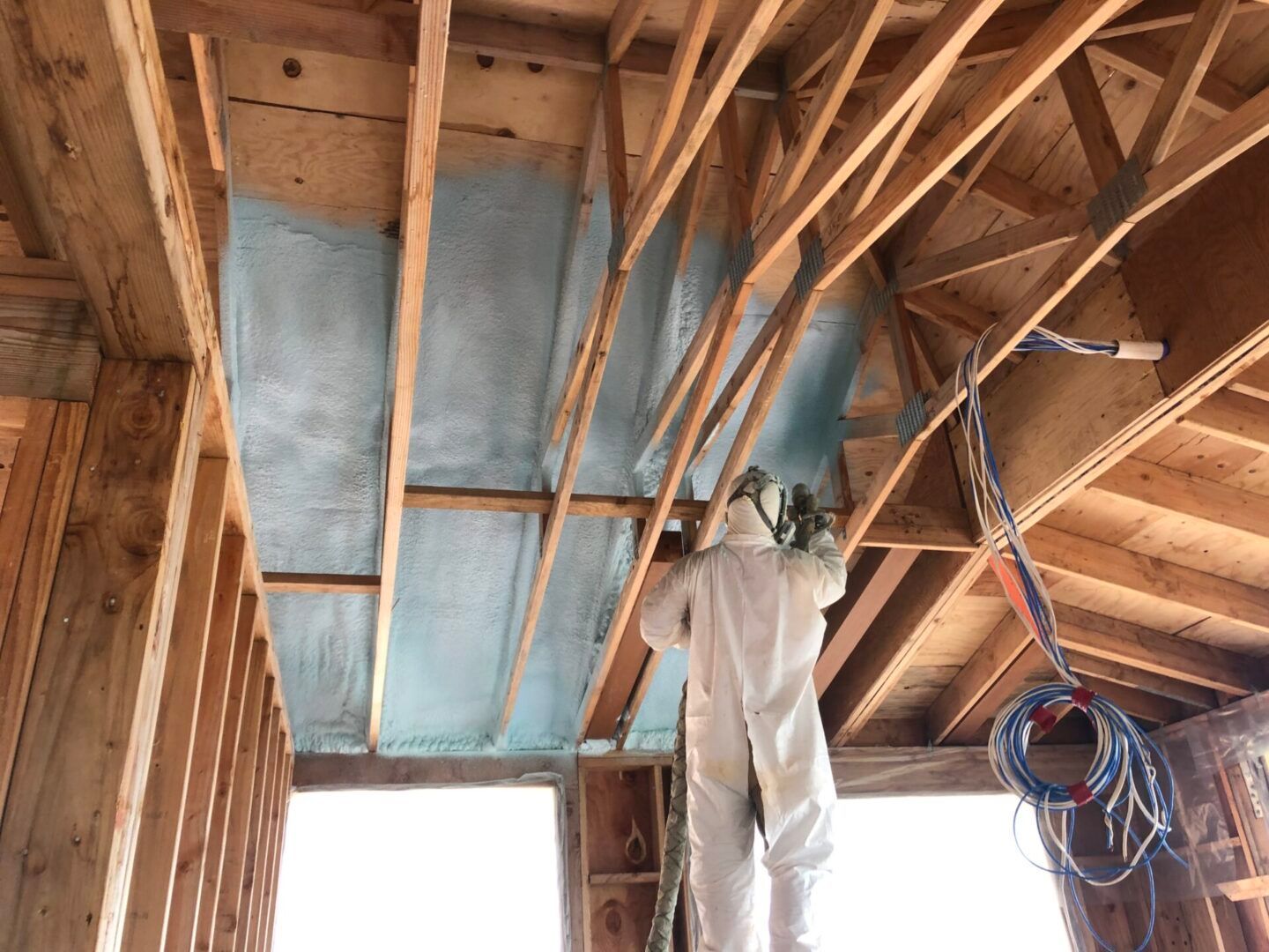 Person in protective suit spraying blue insulation onto wooden beams.
