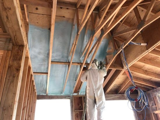 Person in protective suit spraying blue insulation onto wooden beams.