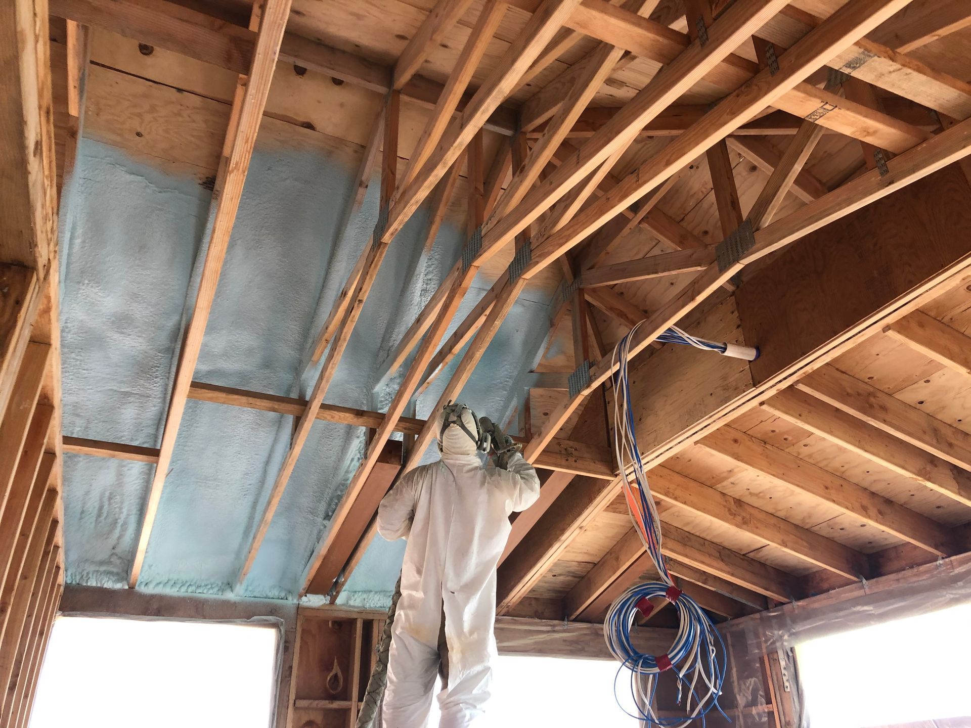 A worker in a protective suit sprays insulation onto a wood ceiling frame in a construction site.
