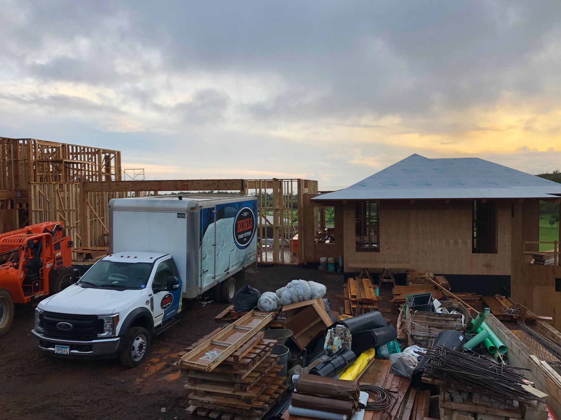 Construction site with wood-framed buildings. White truck and orange lift equipment are present. Cloudy sky overhead.