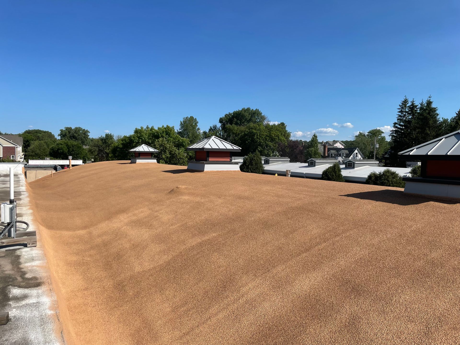 Flat rooftop covered in light brown gravel with several white capped vents, under a blue sky.