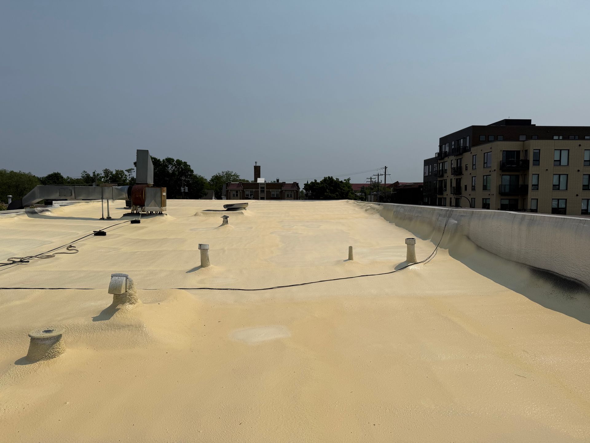 Flat, light-yellow rooftop with vents and pipes, surrounded by low walls, with buildings and sky in the distance.