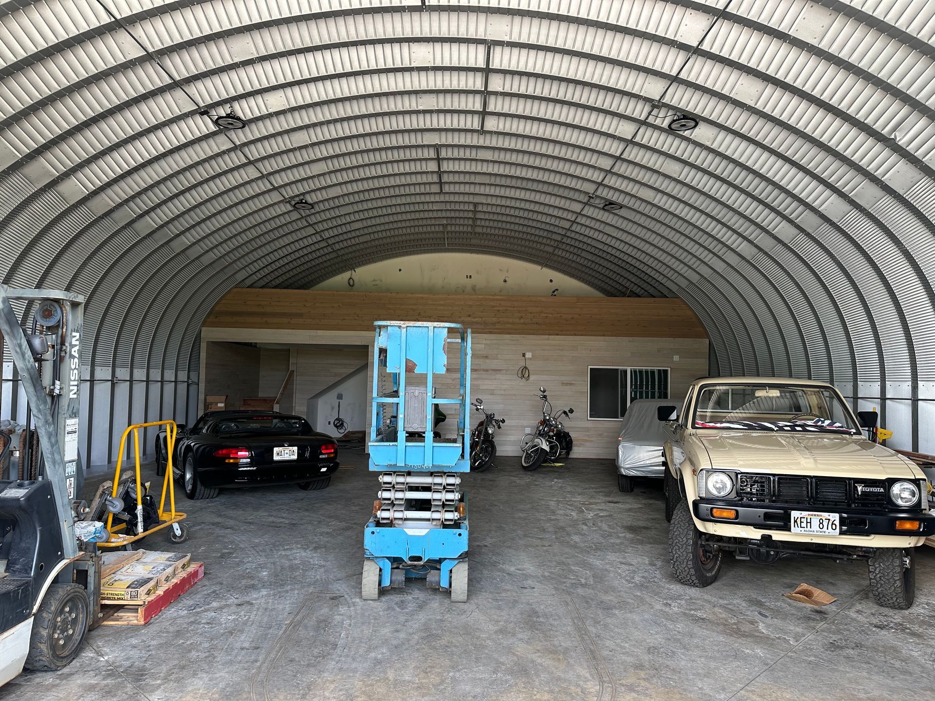 Inside a metal arch building: a blue lift, cars, motorcycles, and a wooden structure.
