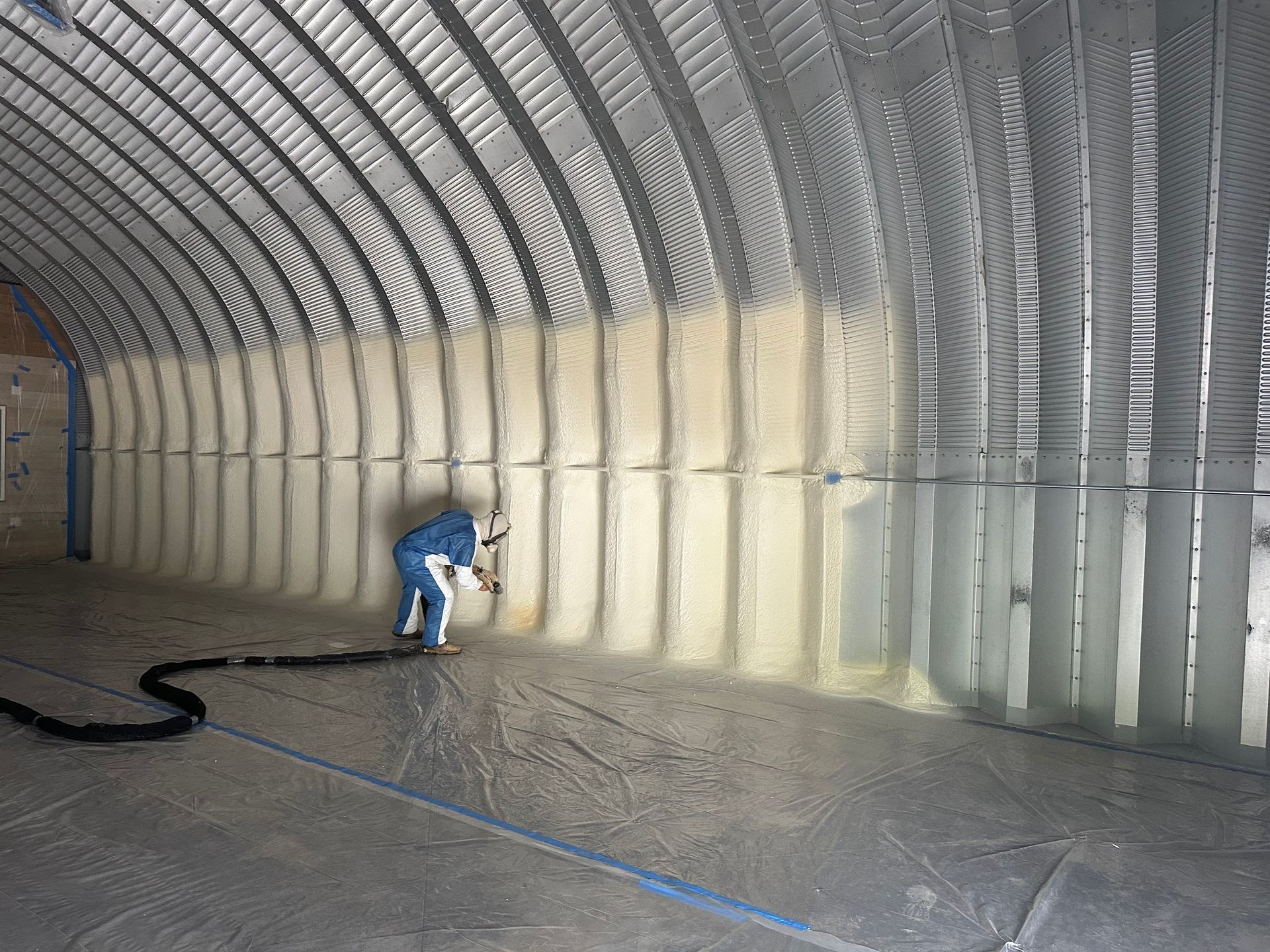 Person spraying insulation inside a large, curved metal structure. The structure has parallel ridges.