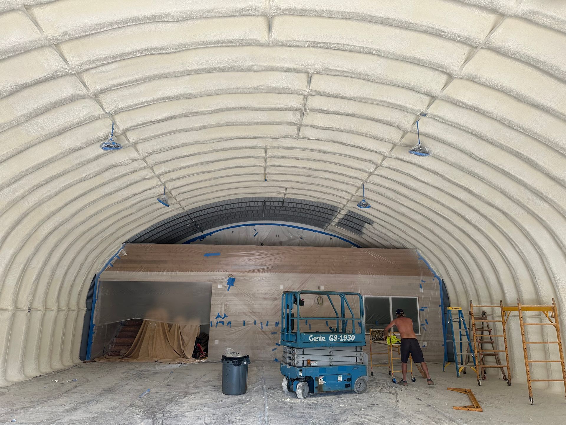 Interior view of a building with arched ceiling and spray foam insulation. A person is working near a lift.
