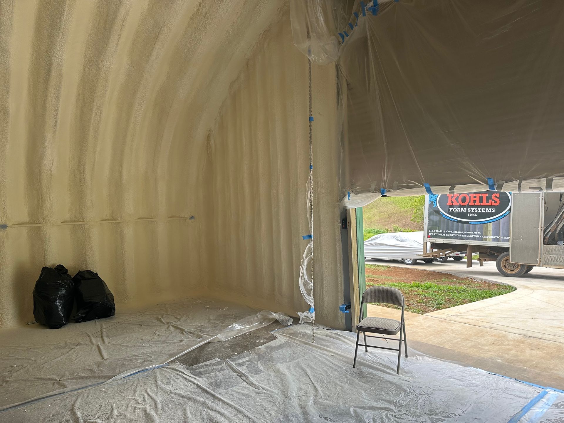 Interior of a Quonset hut being insulated with spray foam. A truck is visible outside.