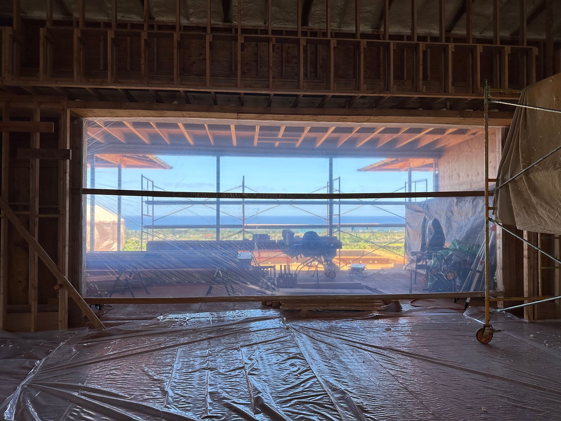 Interior view of unfinished construction with a large window covered in clear plastic, overlooking a landscape.