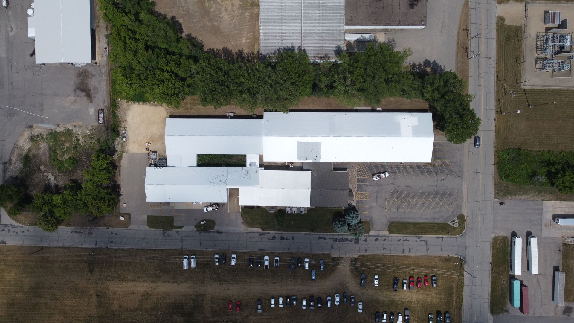Overhead view of several white industrial buildings with trees, parking areas, and surrounding fields.