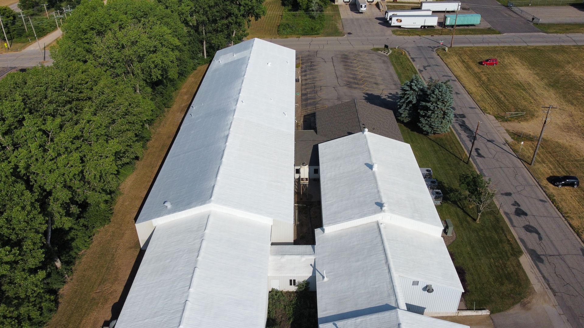 Aerial view of white industrial buildings with angled roofs, next to a paved area with trees and vehicles.
