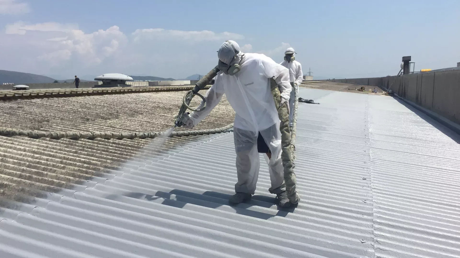 Workers spraying sealant on a corrugated metal roof, wearing protective suits and masks, outdoors on a sunny day.