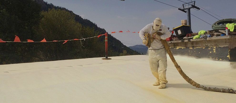 Person in protective gear sprays foam onto a surface, likely a roof, with mountains in the background.