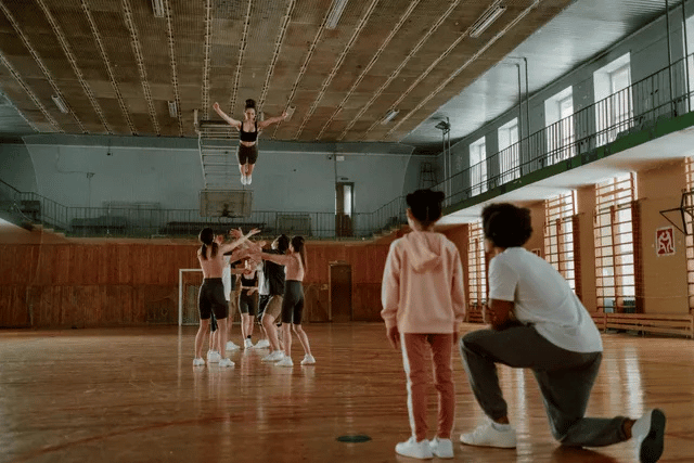 Gymnast in mid-air above teammates. Gymnasium setting; light-colored clothing. Coach kneeling, watching.