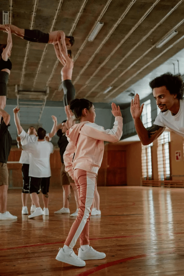 People in gym performing human pyramid, child high-fiving man. Wooden floor, large windows, and high ceiling.