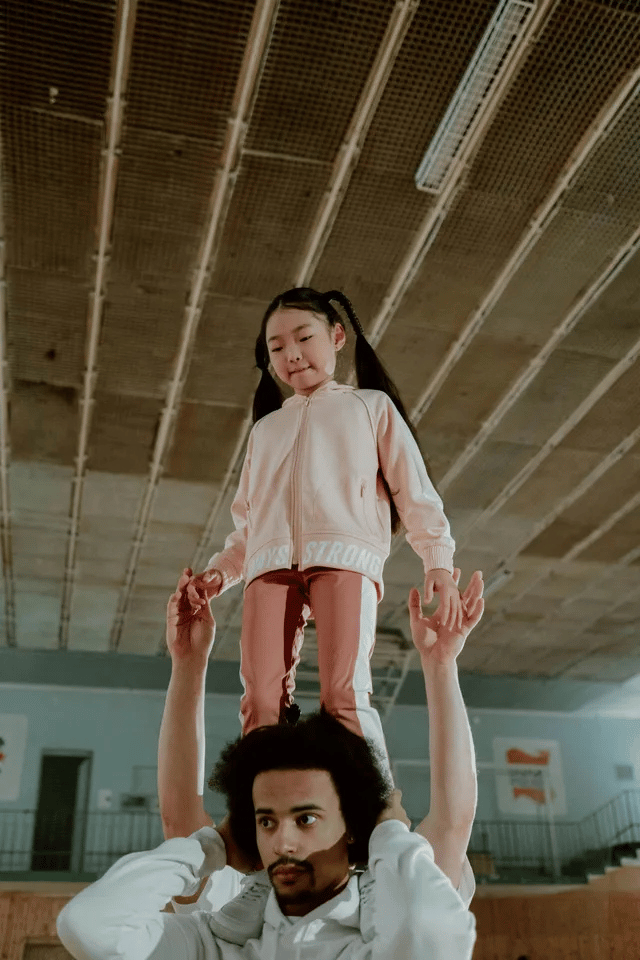 Person holds a child on their shoulders in a gymnasium. The child smiles, wearing pink.