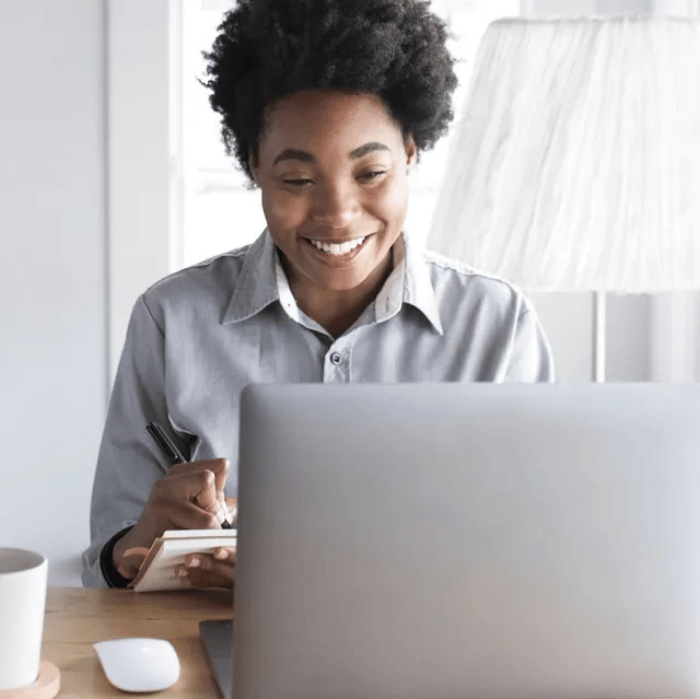 Woman in grey shirt smiling, writing in a notebook while looking at a laptop at a desk.