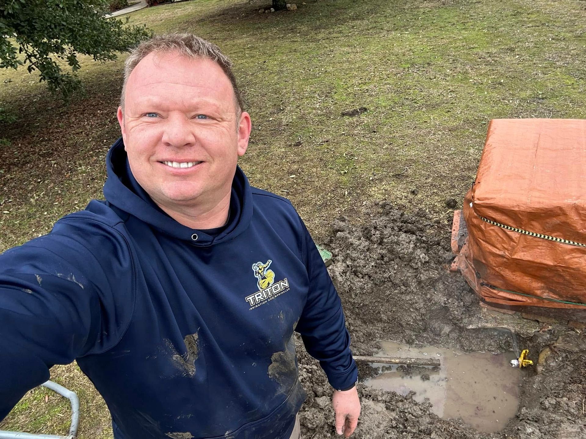 Man in blue hoodie smiles near a muddy excavation, possibly plumbing work.