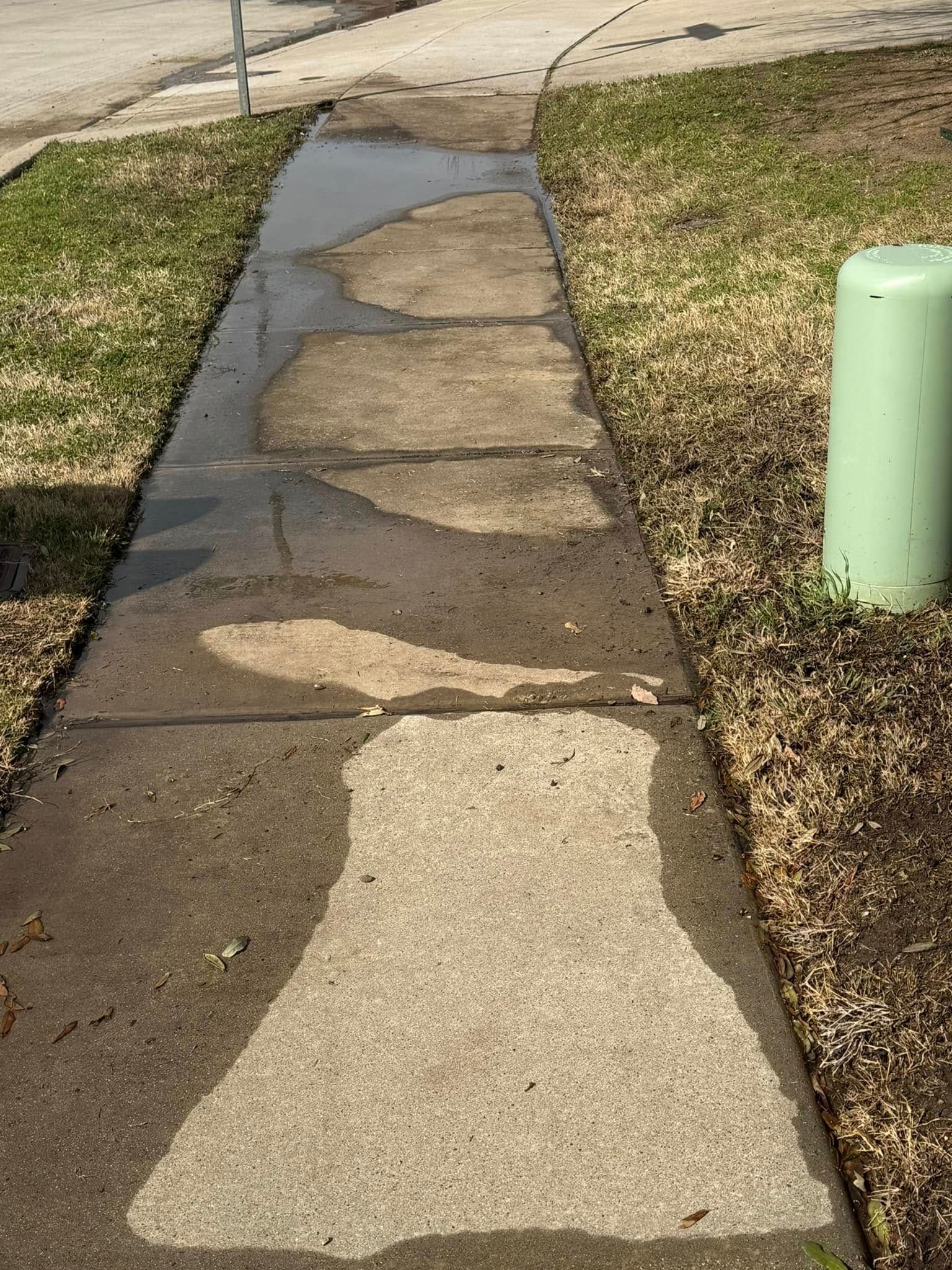 Wet sidewalk with patches of water next to grass and a light green utility box.