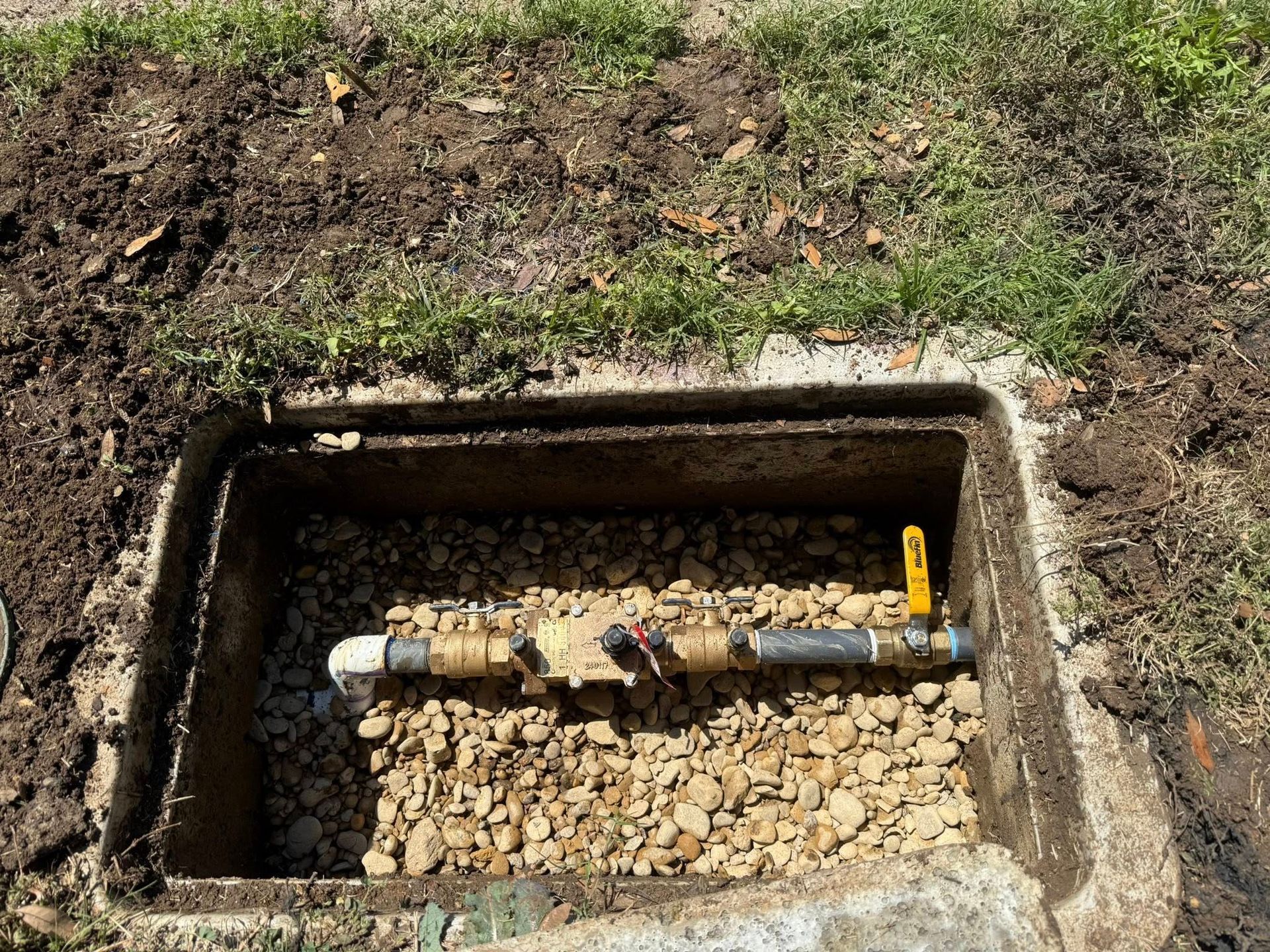 Open rectangular concrete box containing a water valve and pipe, surrounded by gravel.