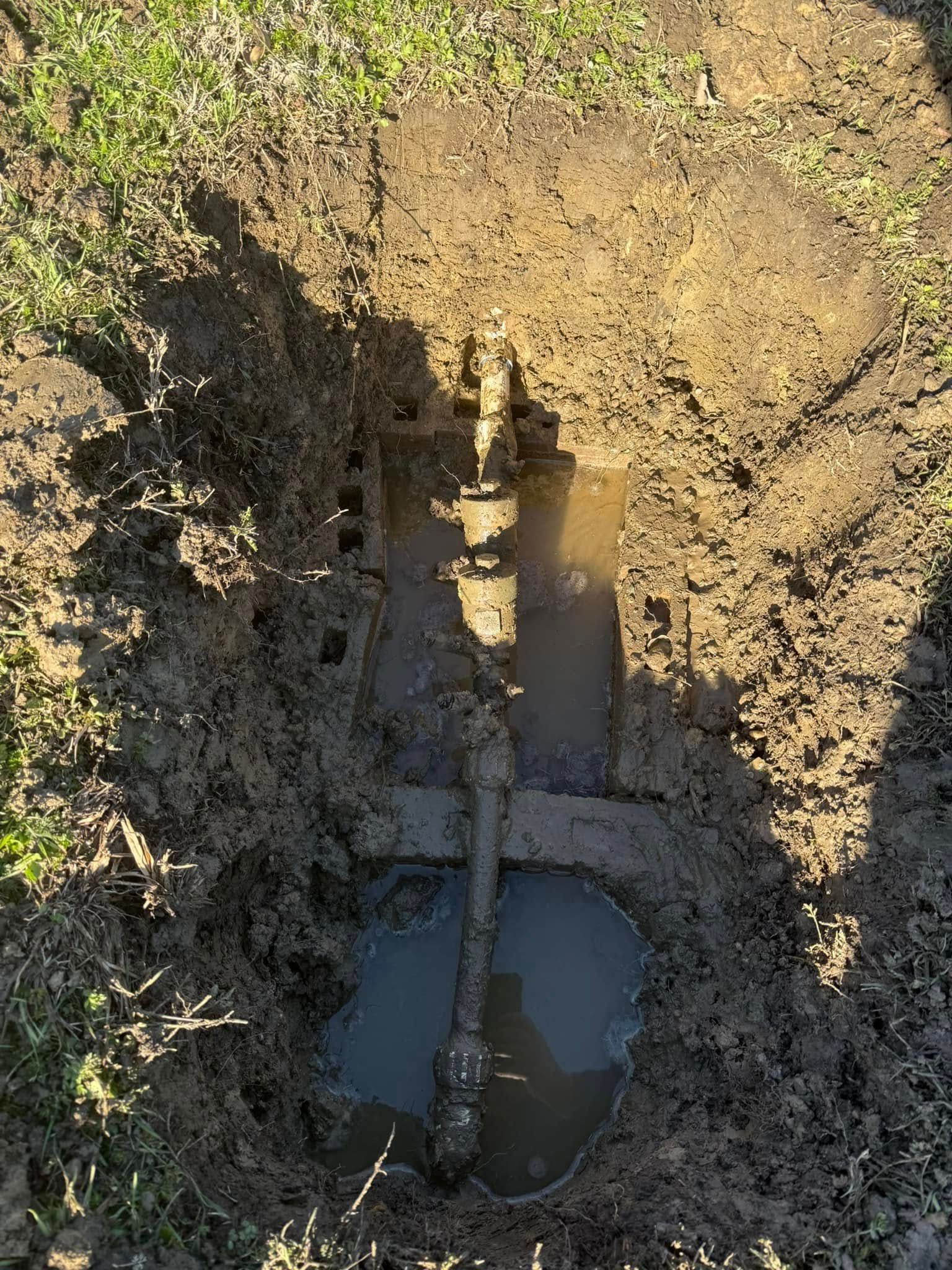 A shovel dug into muddy ground, revealing metal pipes and standing water.