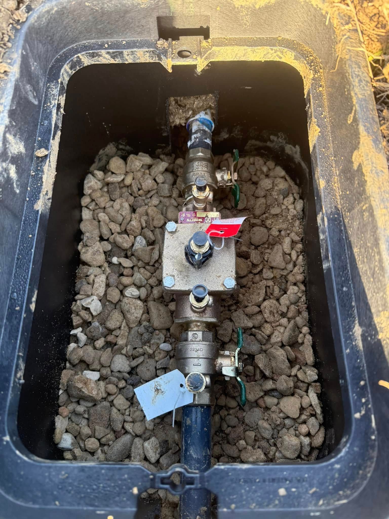 Black plastic box with irrigation valves, gravel, and connected pipes.