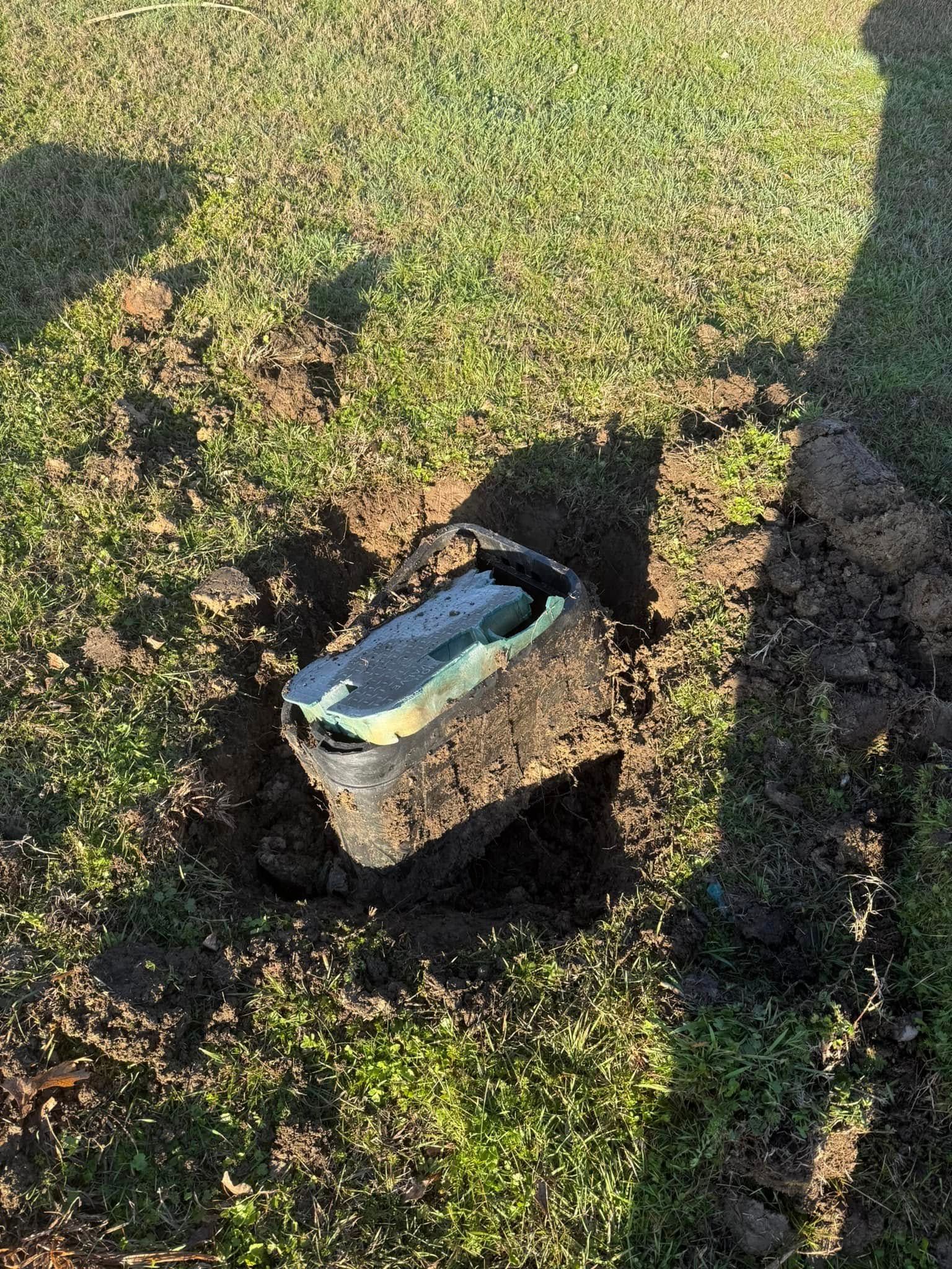 Rectangular, black utility box in a hole in the grass. The box's lid is partially open, revealing green and black inside.
