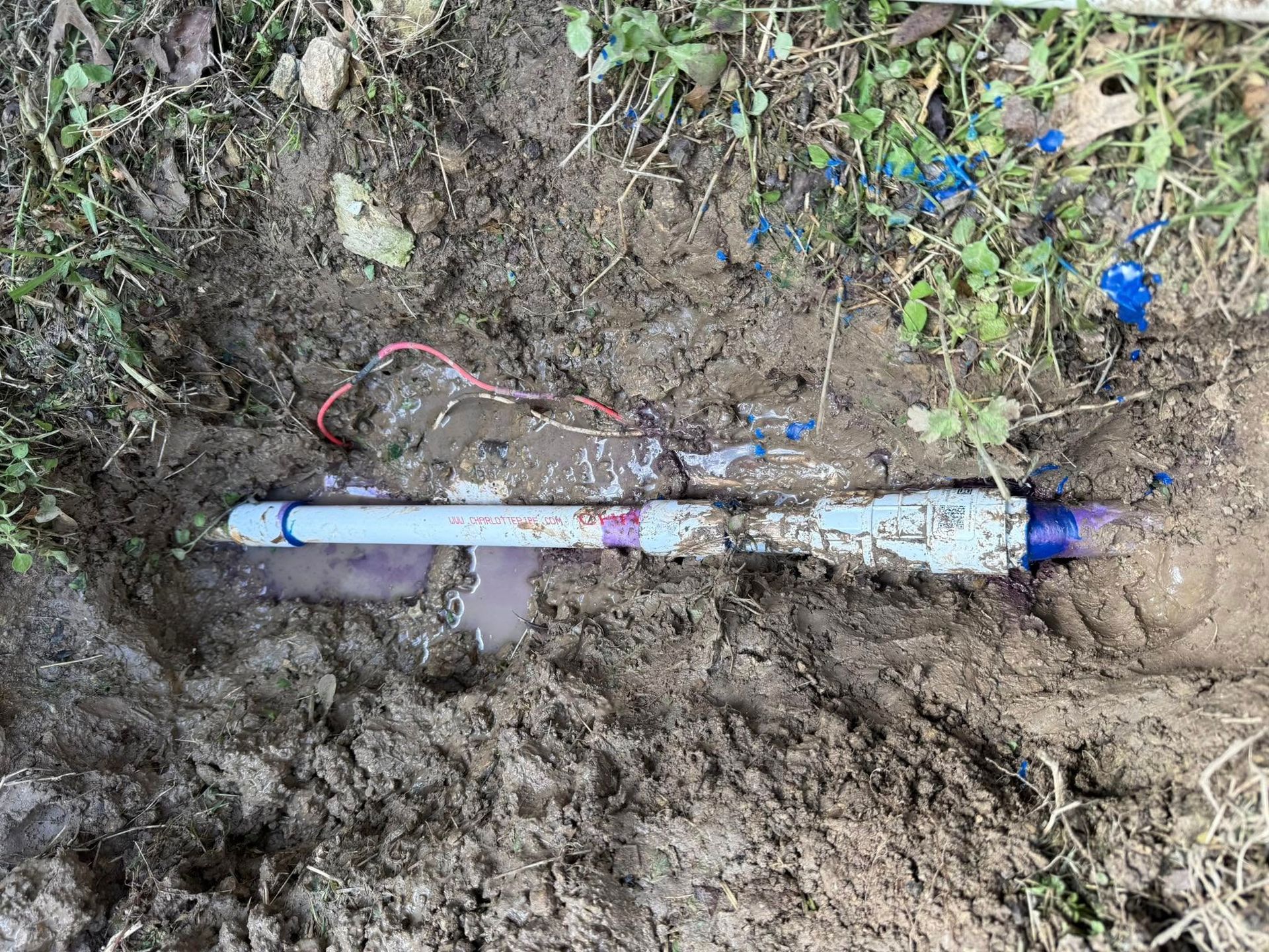 A buried sprinkler valve with white and purple PVC pipes, red and white wires, surrounded by dirt and grass.